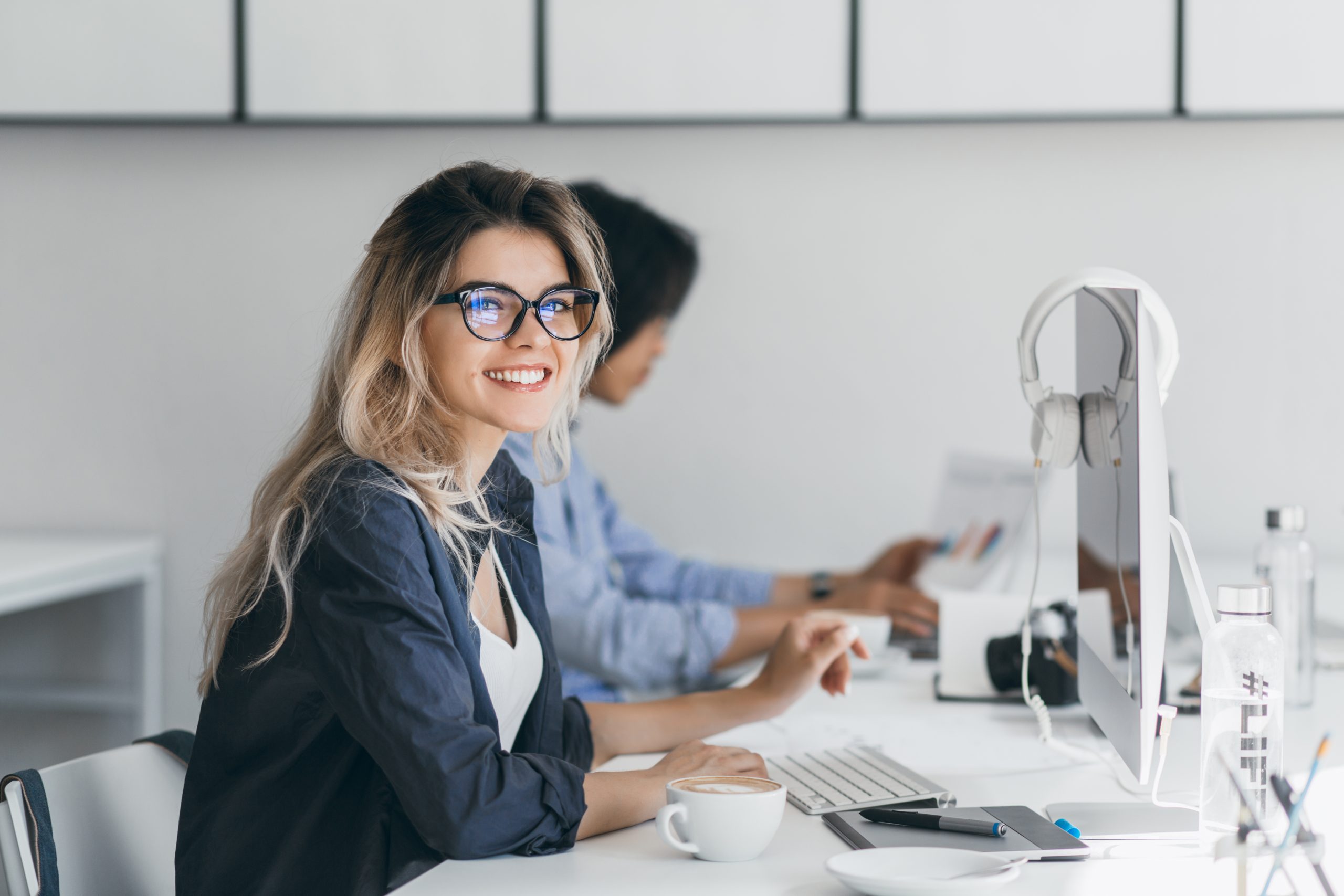 Attractive laughing freelancer girl posing with cup of coffee at her workplace. Chinese student in blue shirt works with document in campus with blonde friend in glasses.